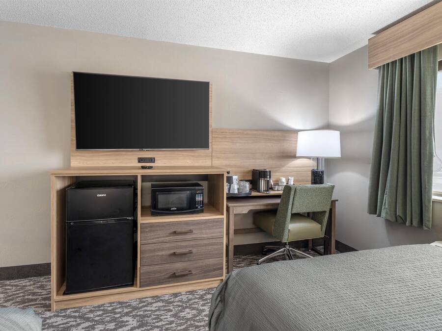 Quarto de hotel com cama, escrivaninha com computador, micro-ondas, frigorífico, televisão de ecrã plano e janela com cortinas.