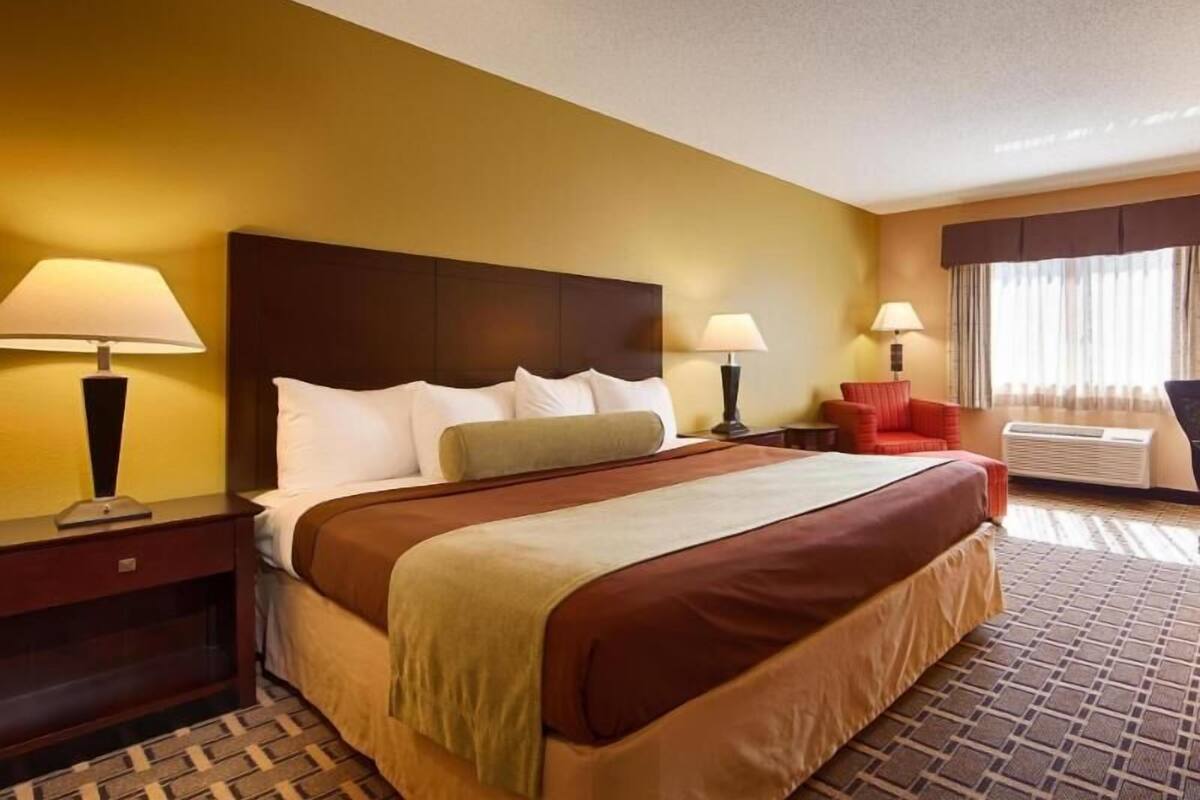 A hotel room with a large bed, two bedside tables with lamps, a red armchair, and a window with curtains.
