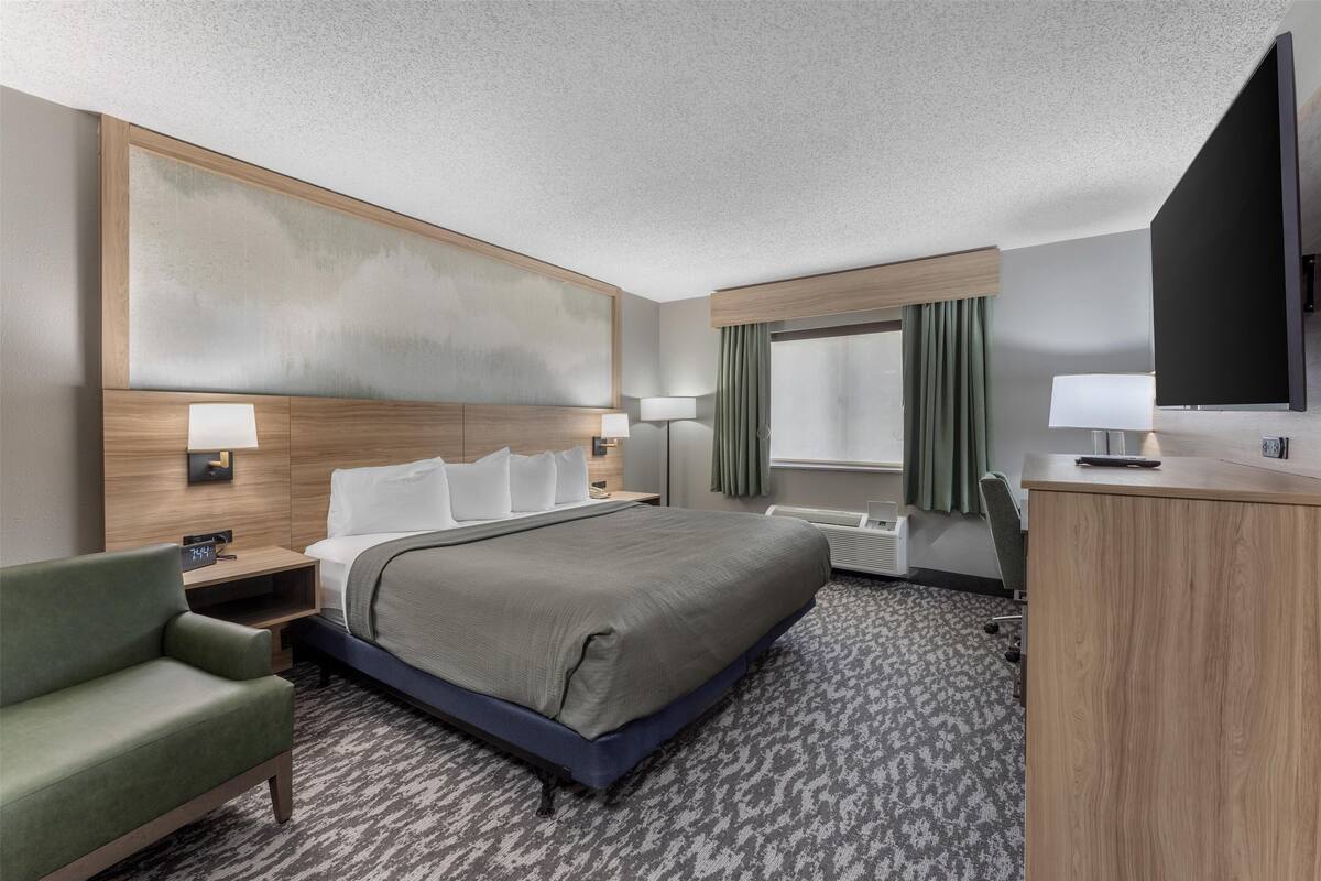 A hotel room with a large bed, a desk, a chair, a flat-screen TV, and a large window with curtains.