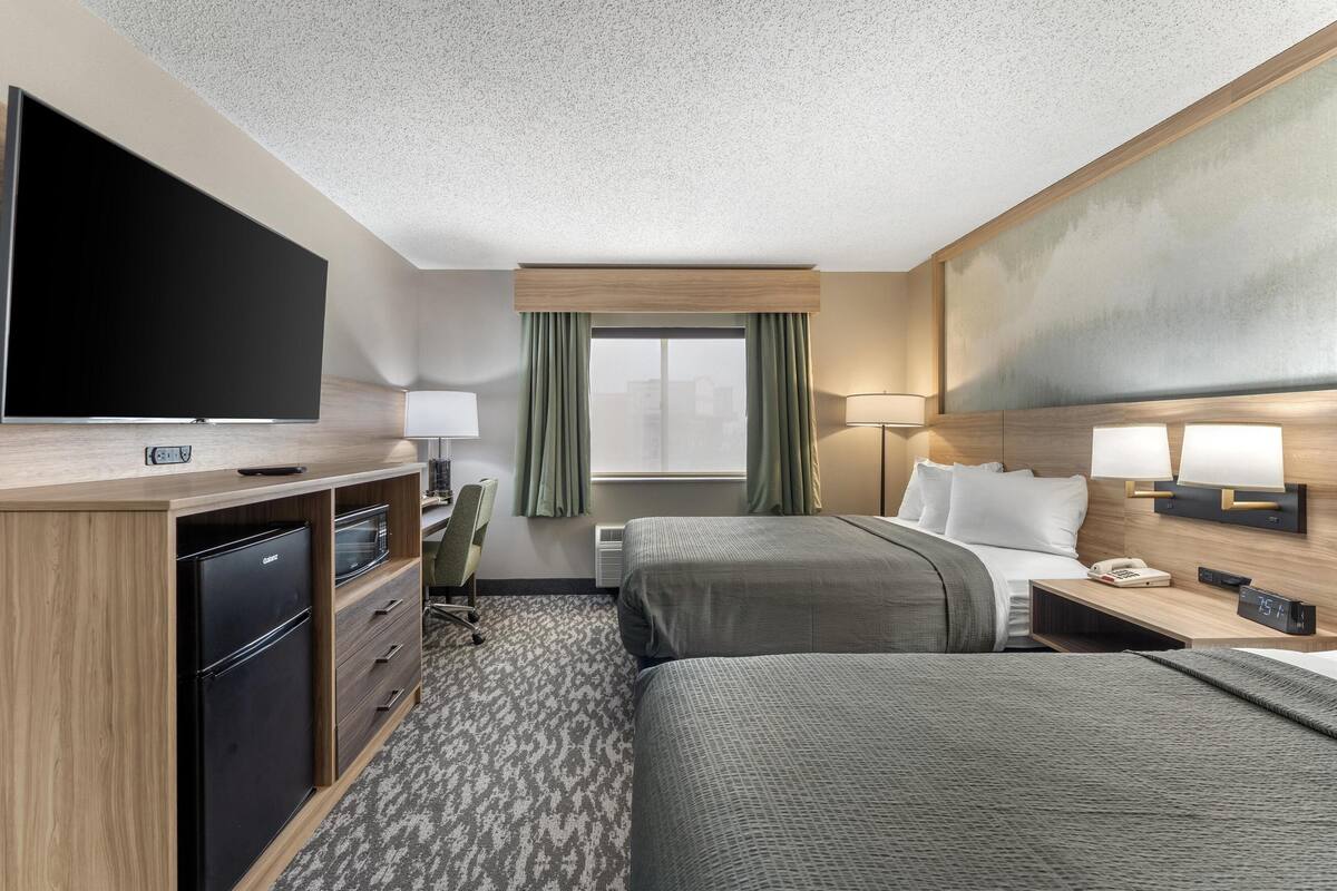 A hotel room with a flat-screen TV, a minibar, a desk with a lamp, two beds, a window with curtains, and a wall-mounted phone.