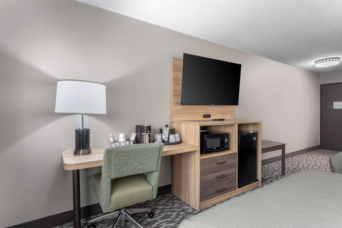 A hotel room with a desk, a flat-screen TV mounted on a wooden cabinet, a microwave, a refrigerator, a lamp, and a chair.