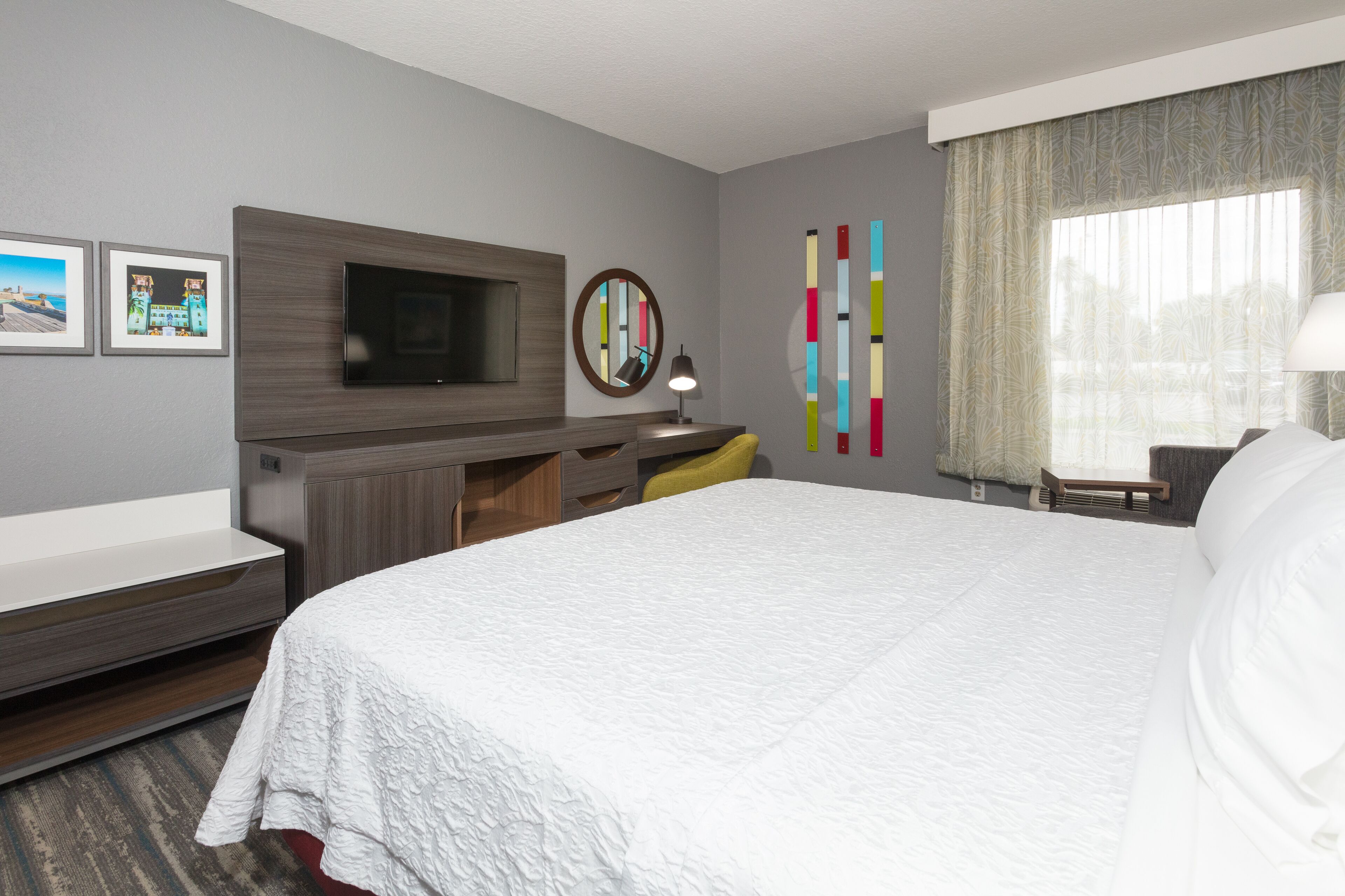 A hotel room with a large bed, a flat-screen TV mounted on a wooden panel, a desk with a chair, a small table with a lamp, and a window with sheer curtains.