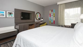 A hotel room with a large bed, a flat-screen TV mounted on a wooden panel, a desk with a chair, a small table with a lamp, and a window with sheer curtains.