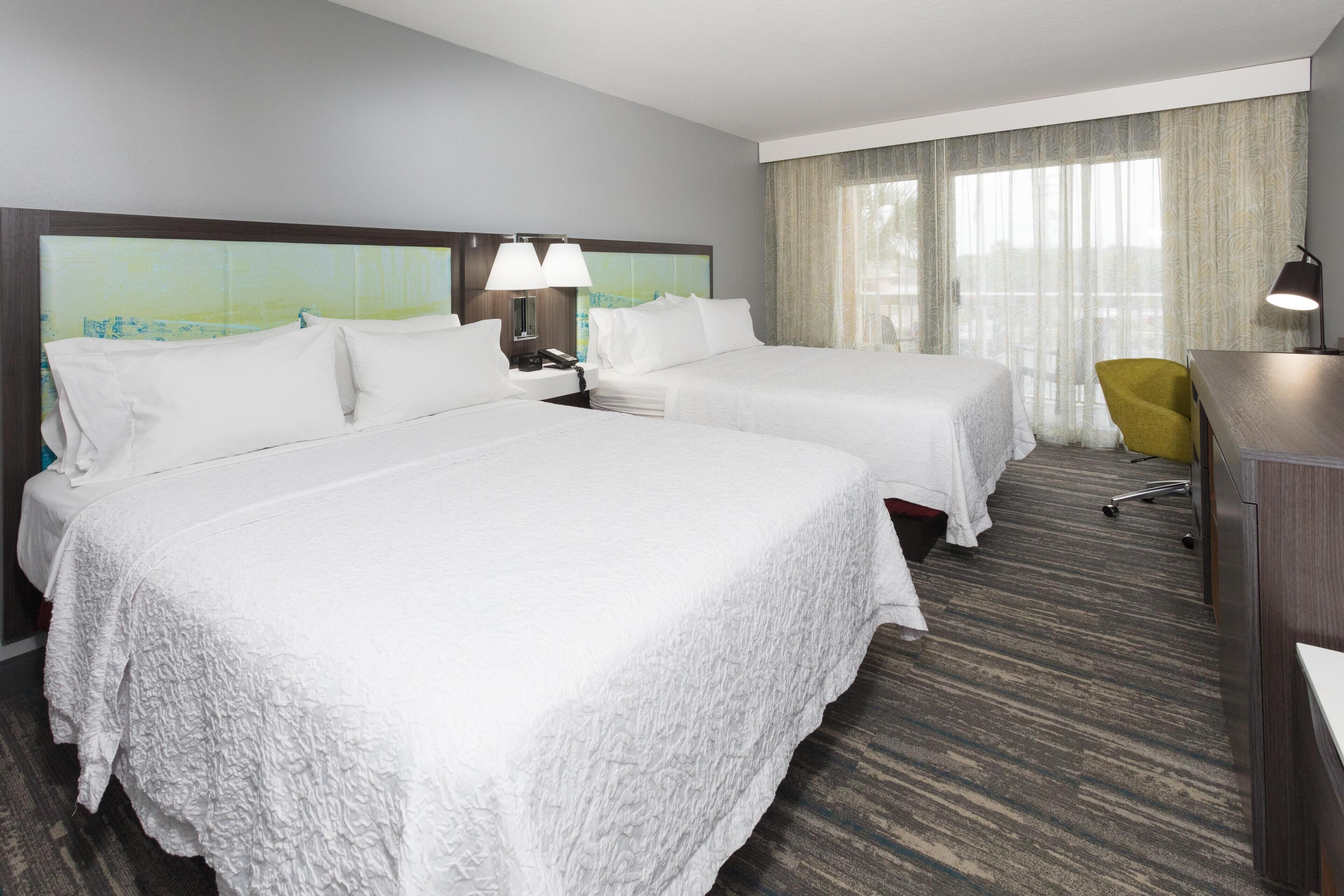 Ein Hotelzimmer mit zwei Betten, einem Schreibtisch, einem Stuhl und einem großen Fenster mit Vorhängen.