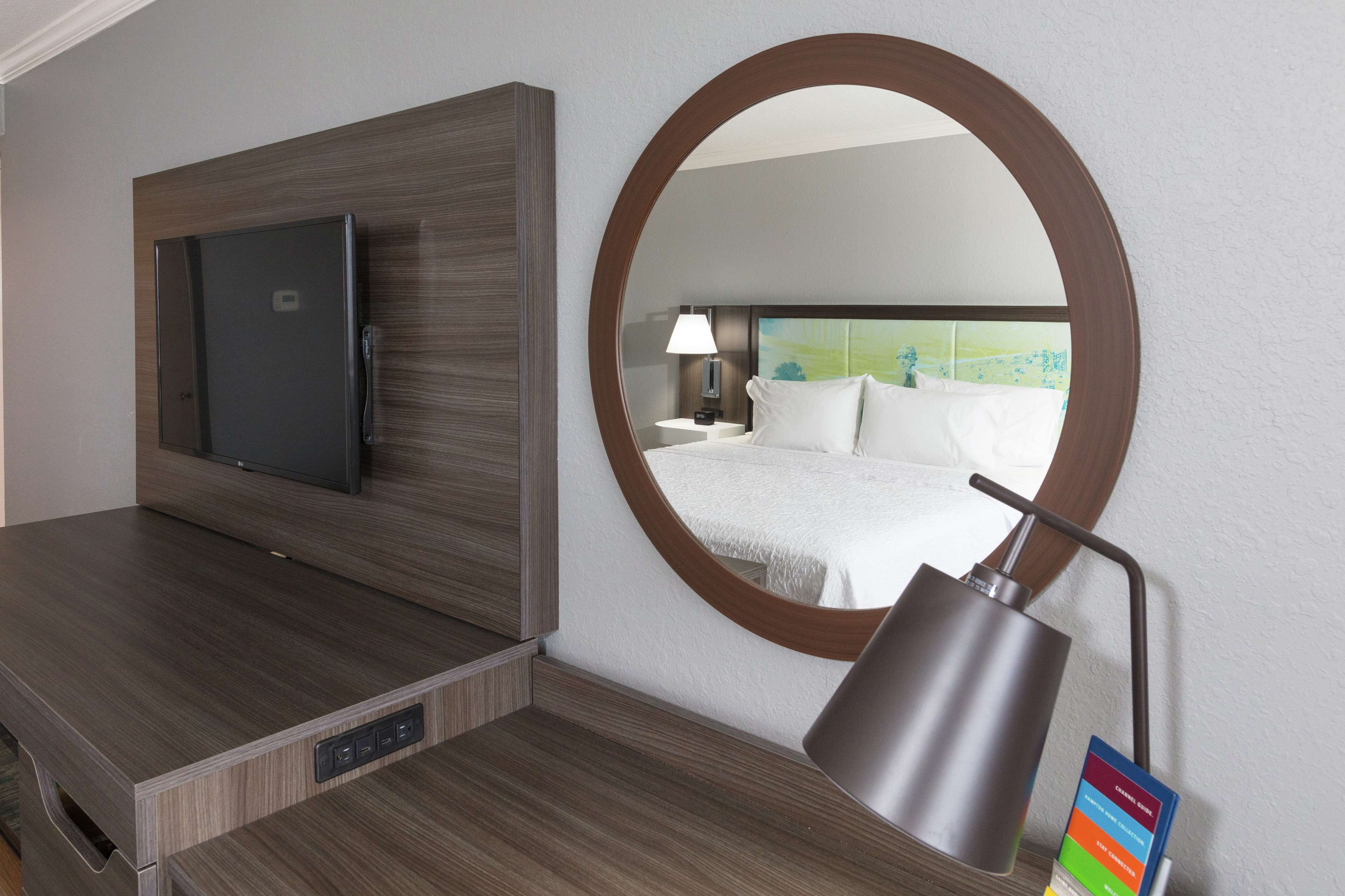 A hotel room with a flat-screen TV mounted on a wooden panel, a round mirror on the wall, and a bedside table with a lamp and a color chart.