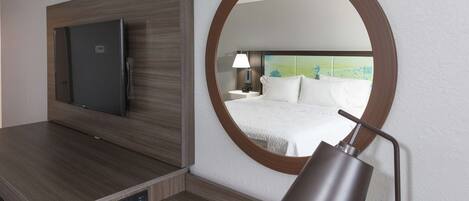 A hotel room with a flat-screen TV mounted on a wooden panel, a round mirror on the wall, and a bedside table with a lamp and a color chart.
