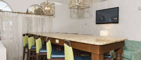 A dining area with a long wooden table, four chairs with yellow and blue cushions, and two hanging light fixtures.