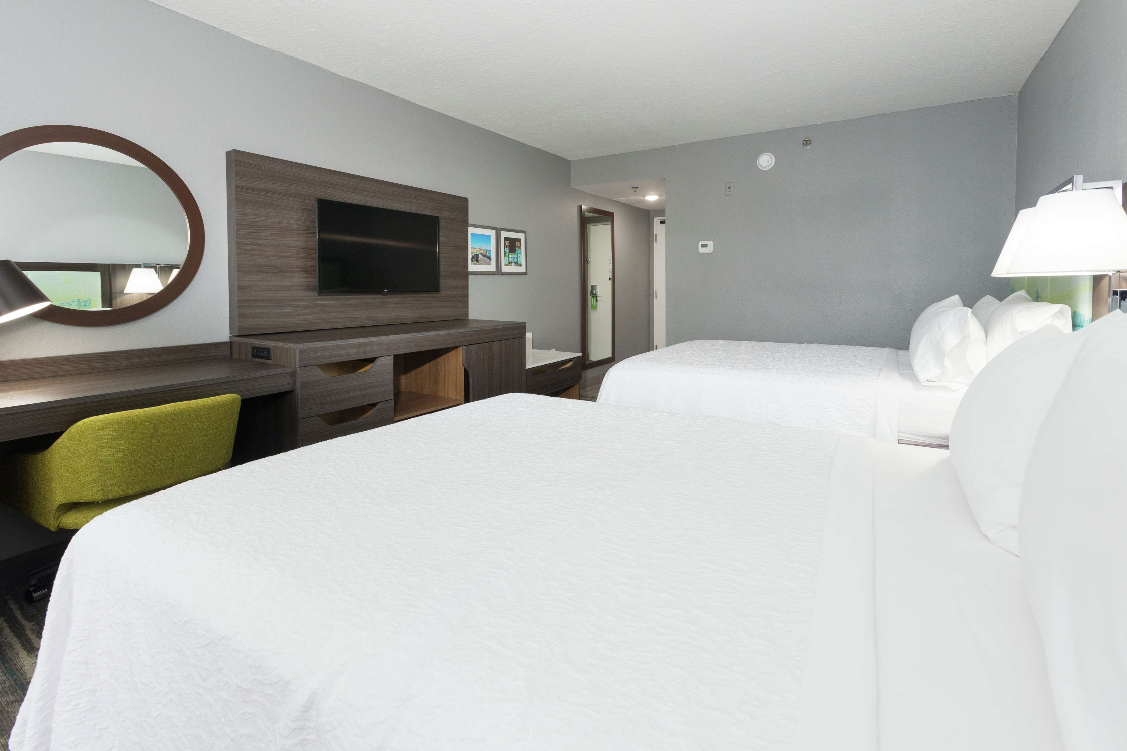 A hotel room with two beds, a desk, a television, a mirror, and a lamp.