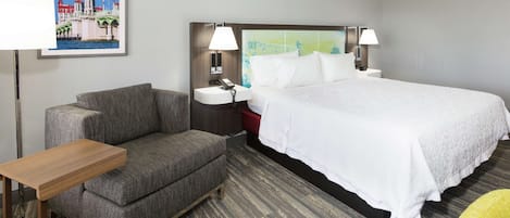 A hotel room with a bed, a chair, a small table, a nightstand, a lamp, and a framed picture on the wall.