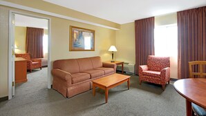 A hotel room with a sofa, armchair, dining table, and a painting on the wall.