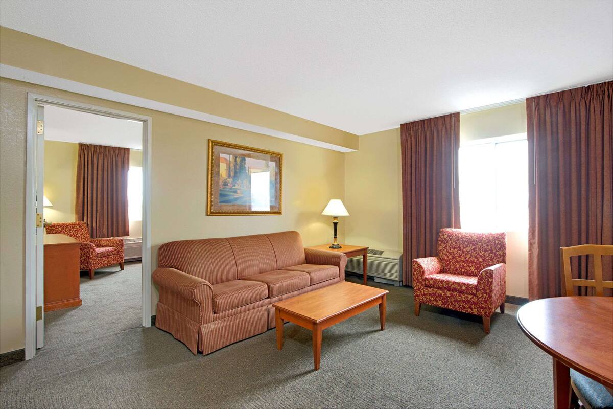 A hotel room with a sofa, armchair, dining table, and a painting on the wall.