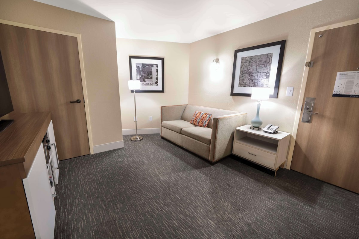 Ein Hotelzimmer mit einer Couch, einem Nachttisch mit Lampe, einer gerahmten Karte an der Wand und einer Tür mit Holzeinlage.