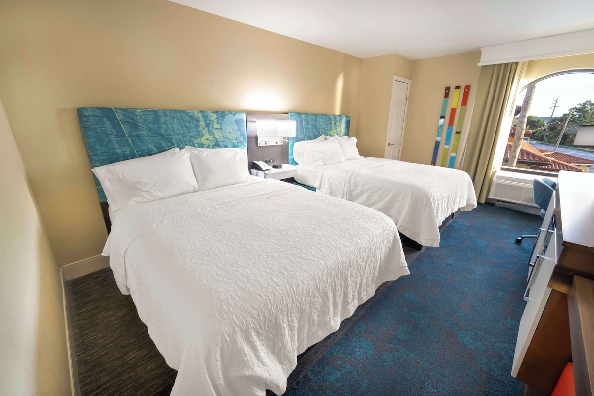 Ein Hotelzimmer mit zwei Betten, einem Schreibtisch, einem Stuhl, einem Nachttisch und einem Fenster mit Vorhängen.