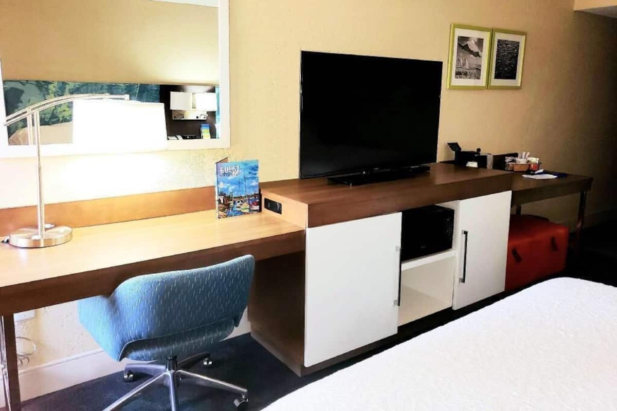 Ein Hotelzimmer mit einem Bett, einem Schreibtisch mit Fernseher, einem Sessel, einer Lampe und einem kleinen Tisch mit einem roten Koffer.