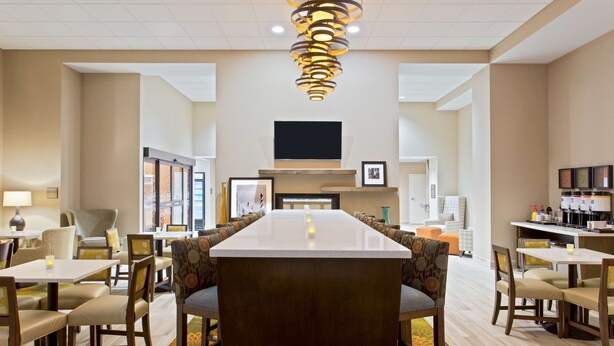 A hotel dining area with a long table, multiple chairs, a modern chandelier, and a flat-screen TV on the wall. - Hampton Inn Sault Ste Marie (Sault Ste. Marie)