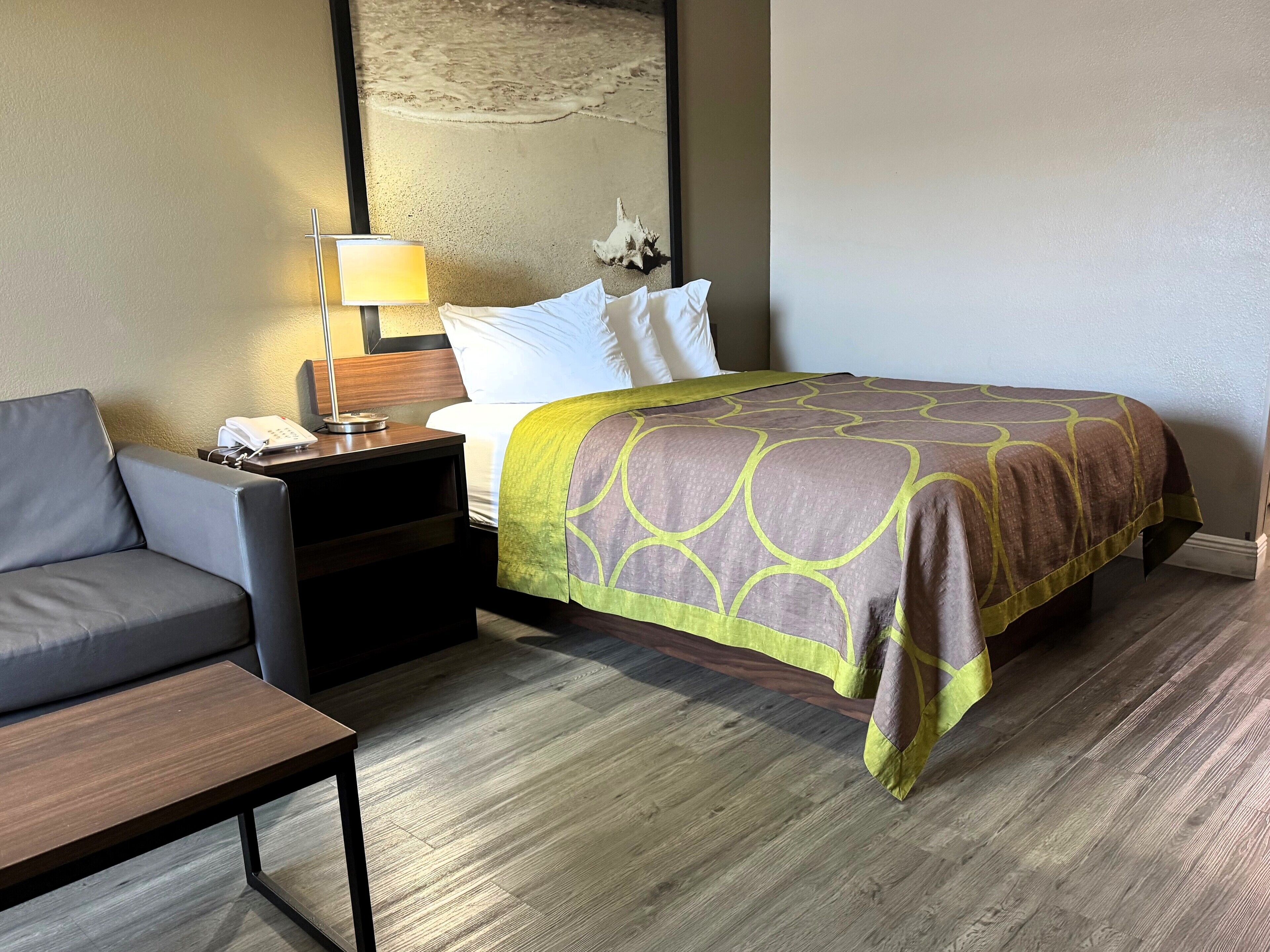 A hotel room with a bed, a desk, a chair, and a nightstand with a lamp.