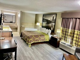 Habitación de hotel con cama, mesitas de noche, escritorio, silla, espejo, televisor montado en la pared, aire acondicionado y un amplio ventanal con cortinas.