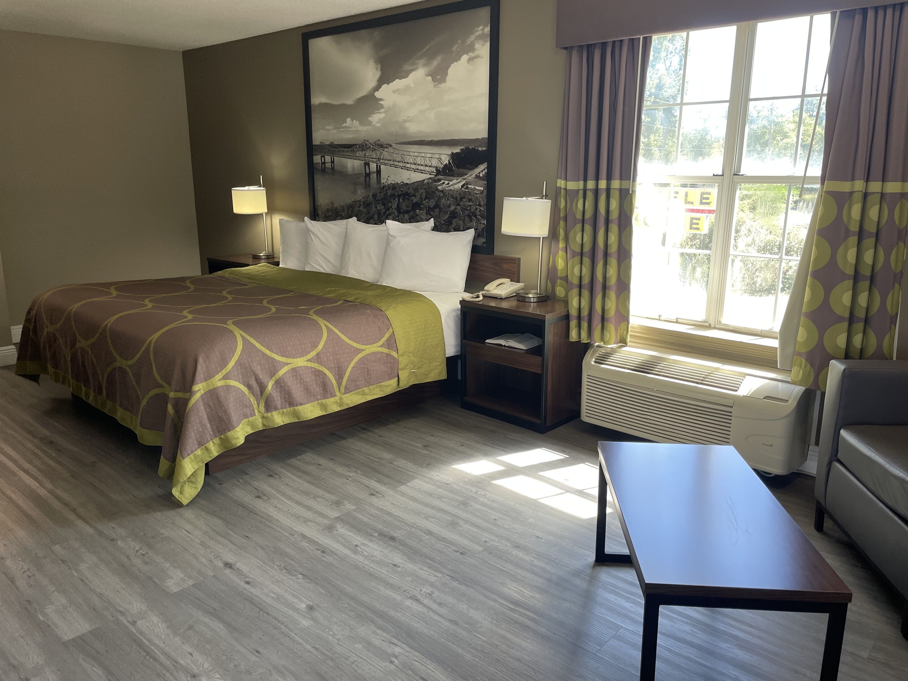 A hotel room with a bed, bedside tables, a desk, a chair, a large window with curtains, and a wall-mounted air conditioner.
