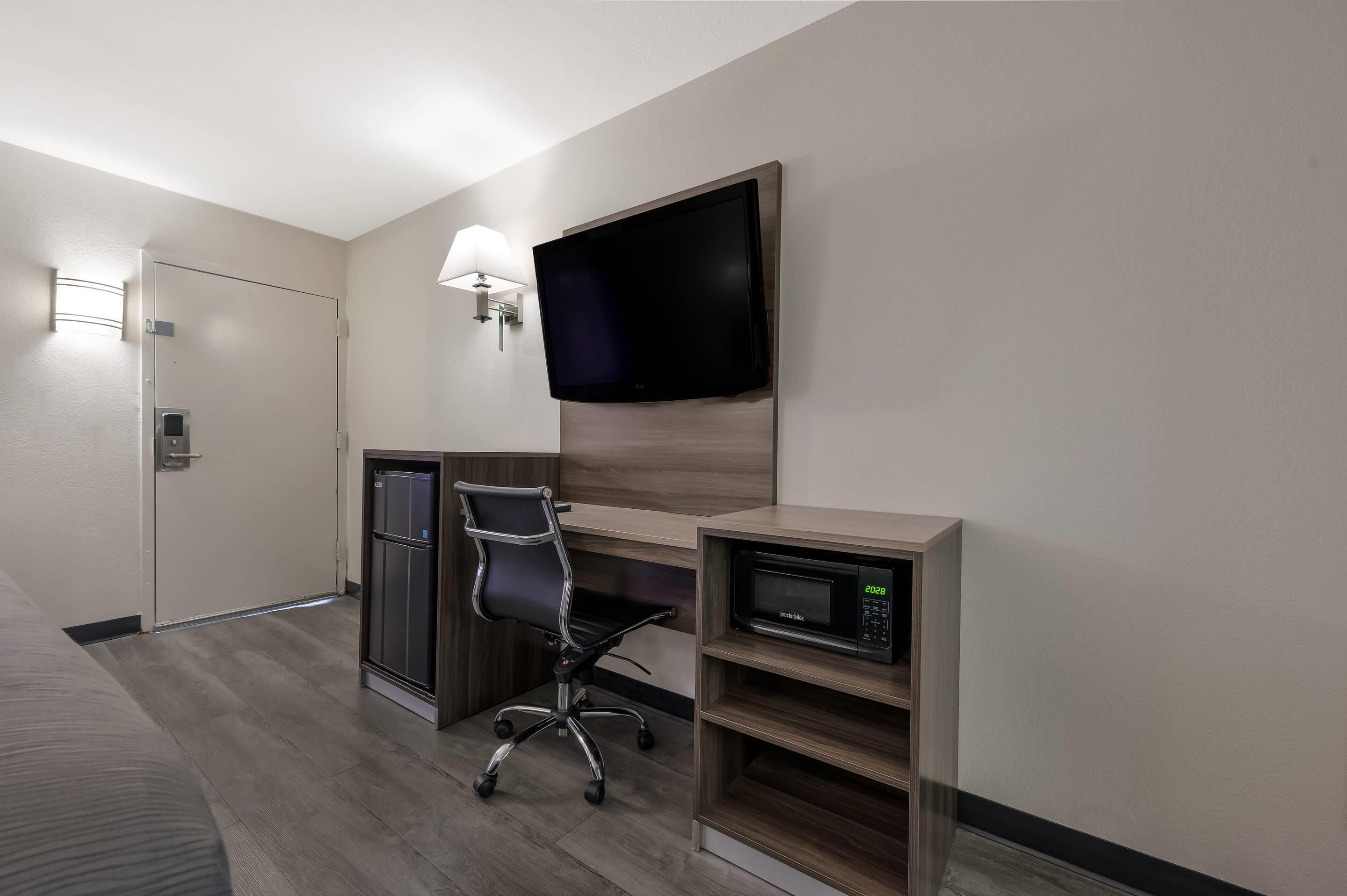Quarto de hotel com uma cama, uma mesa com cadeira, uma televisão na parede, micro-ondas e uma porta com fechadura.