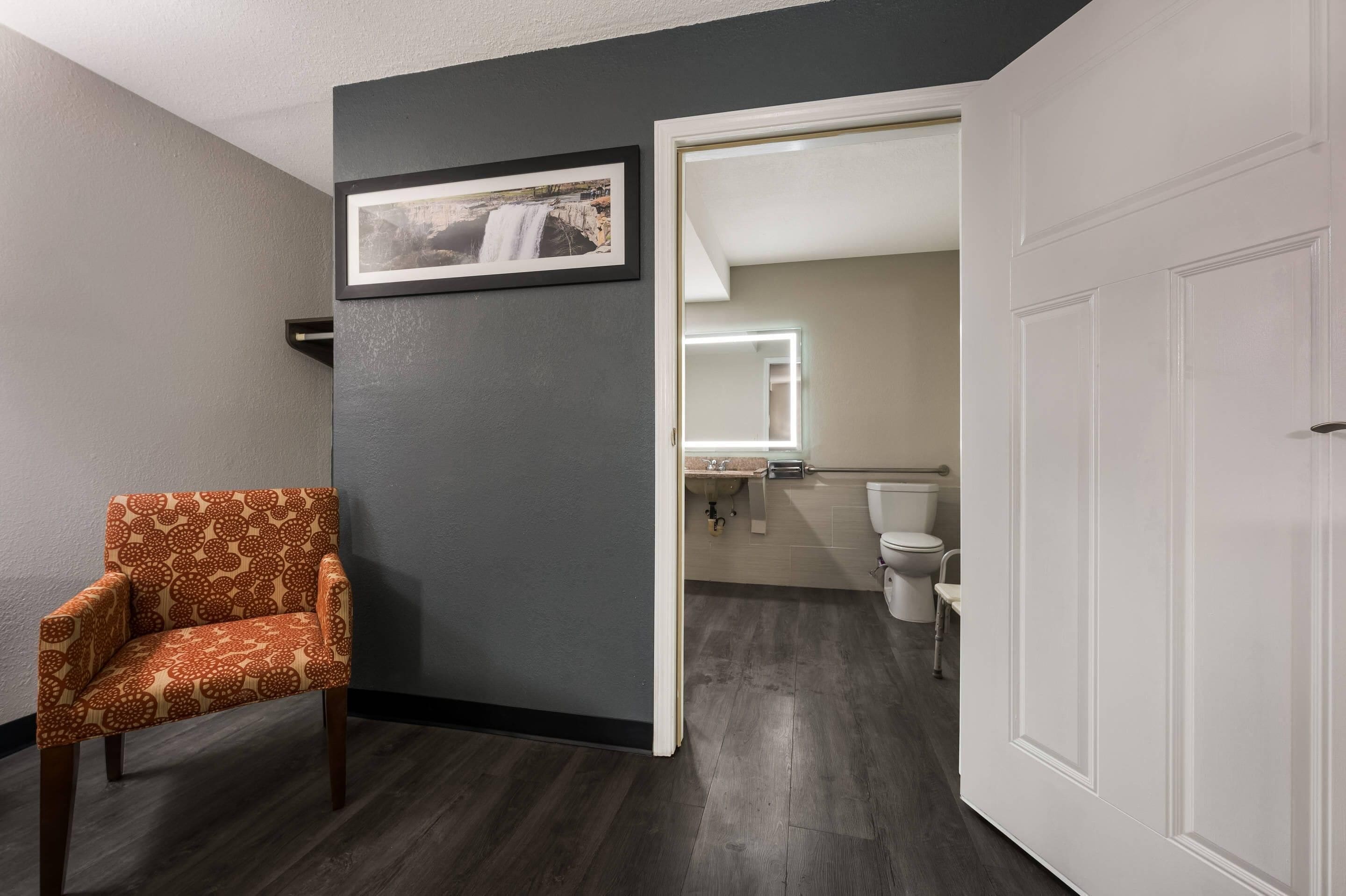 A room with a patterned armchair, a framed picture on the wall, and a doorway leading to a bathroom with a toilet and sink.