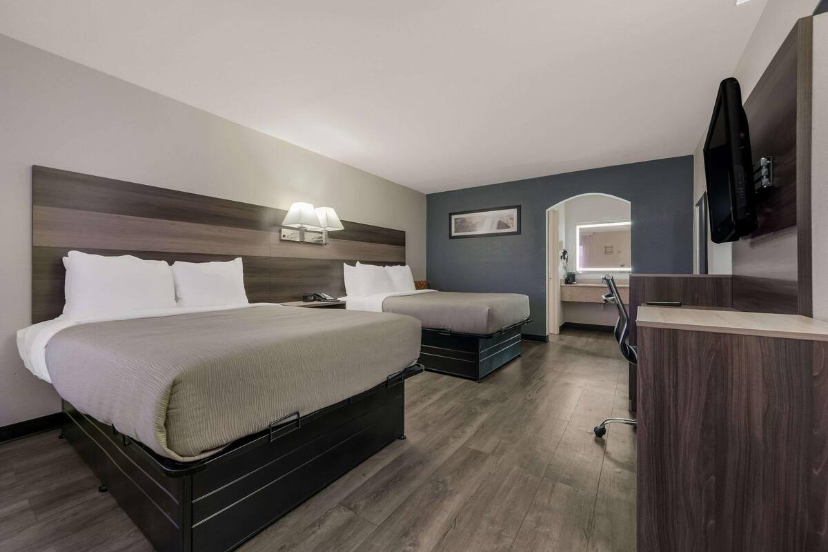 A hotel room with a large bed, a smaller bed, a desk with a chair, a TV, and a bathroom with a mirror.