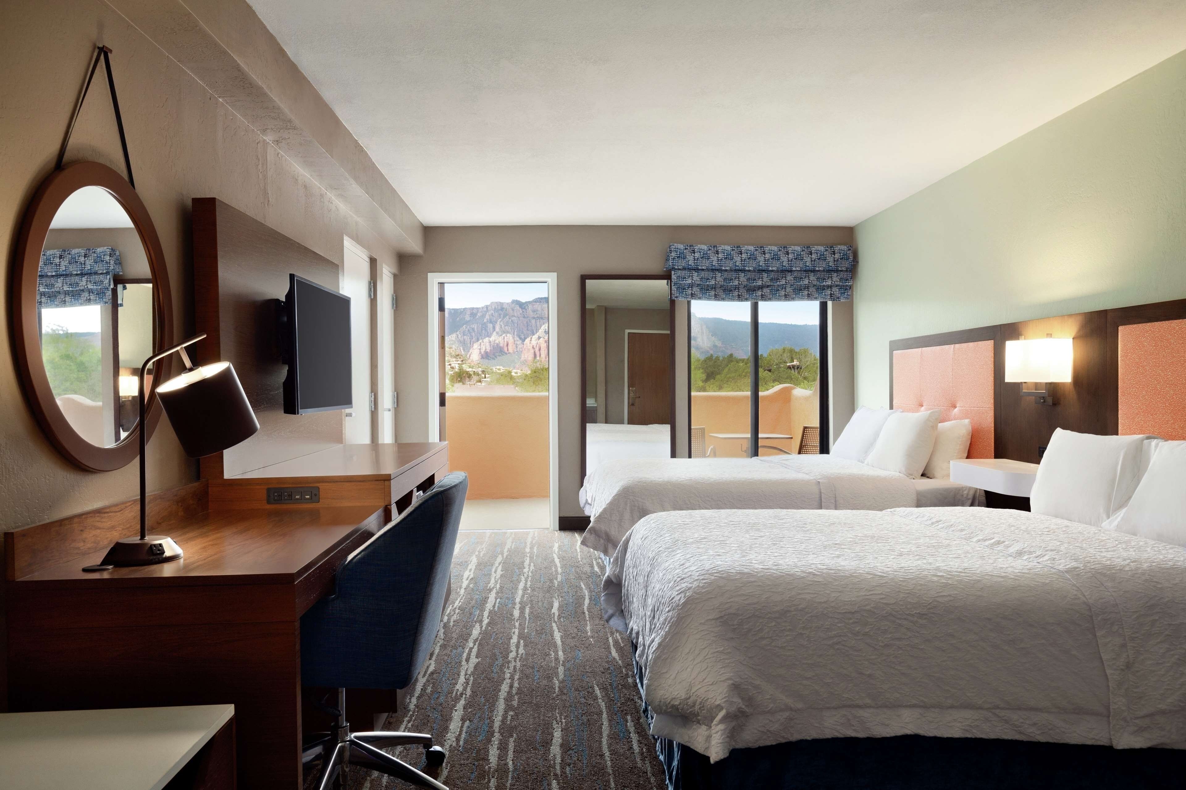 A hotel room with a desk, two beds, a TV, and a large window with a mountain view.