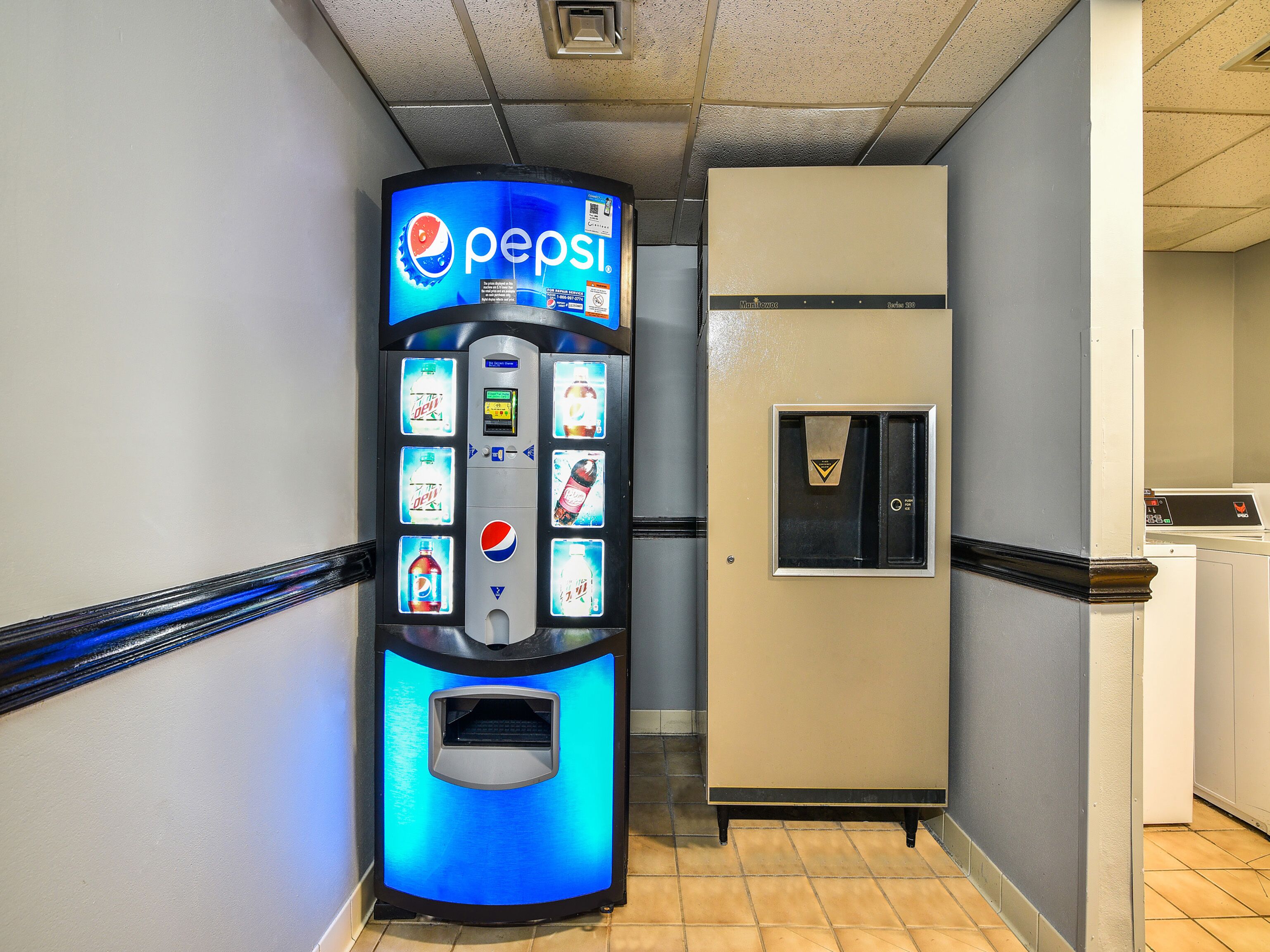 A vending machine with Pepsi branding and a refrigerator in a hallway.