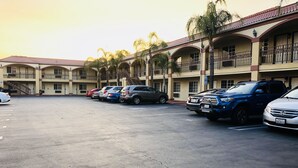 A parking lot with several cars, a building with multiple balconies and a red-tiled roof, and palm trees. - Dynasty Suites Hotel Riverside (Riverside)