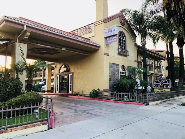 A hotel entrance with a sign for Dynasty Suites, a covered entrance, and a gated driveway.