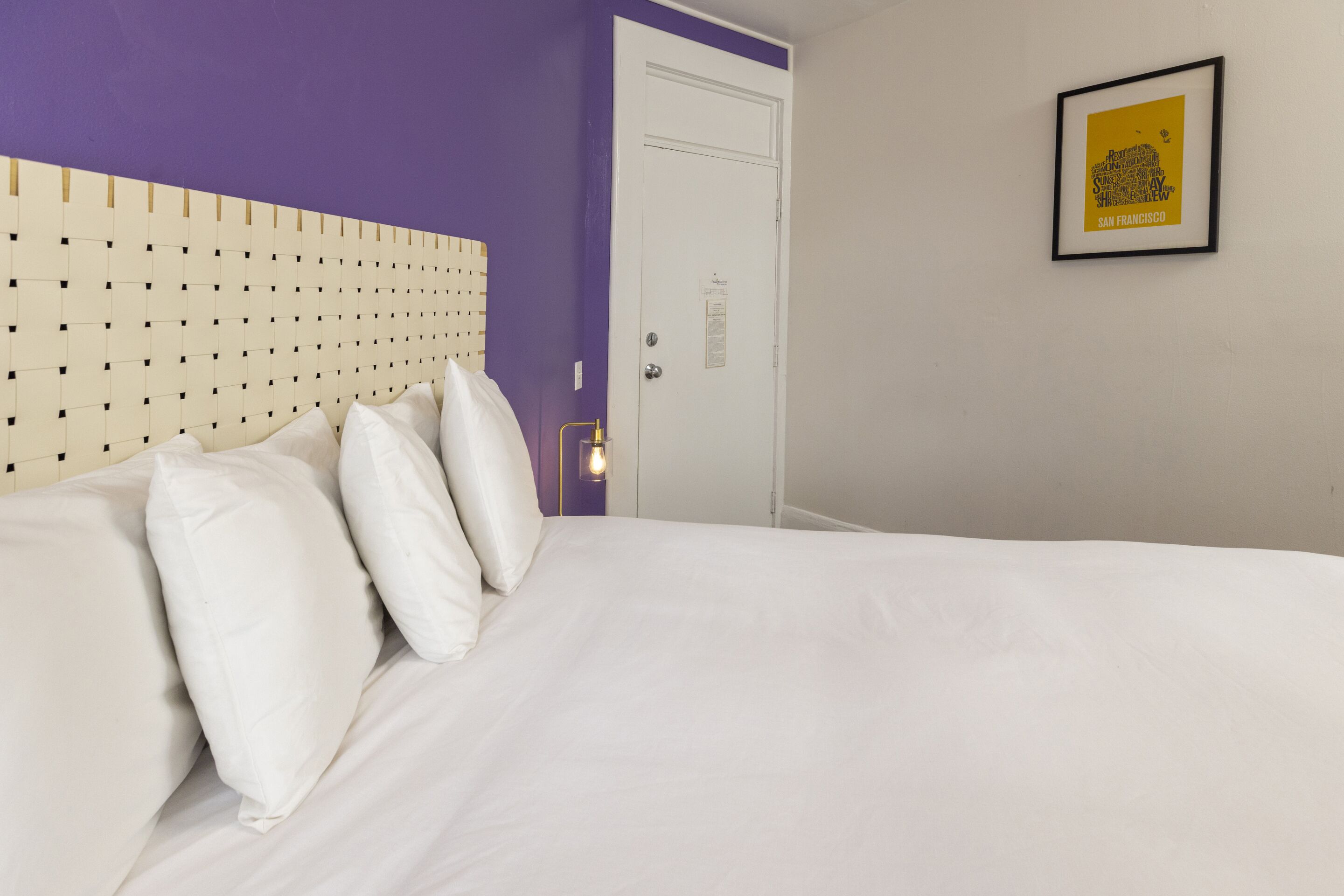 Een hotelkamer met een bed, wit beddengoed, een hoofdeinde met een ruitpatroon, een deur en een lijst met een afbeelding aan de muur.