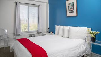 A hotel room with a large bed, a red throw blanket, a clear chair, a bedside table with a clock, and a framed picture on the wall.