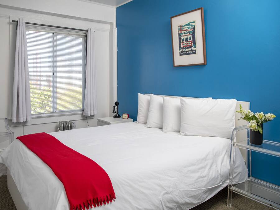 A hotel room with a large bed, a red throw blanket, a clear chair, a bedside table with a clock, and a framed picture on the wall.