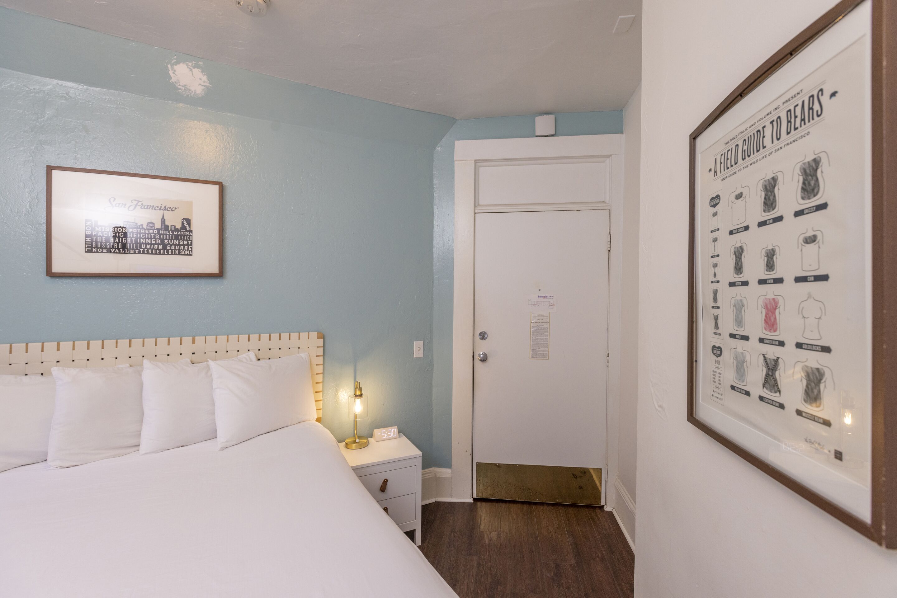 Una habitació d'hotel amb un llit, una taula de nit, un quadre amb una vista urbana i una porta.