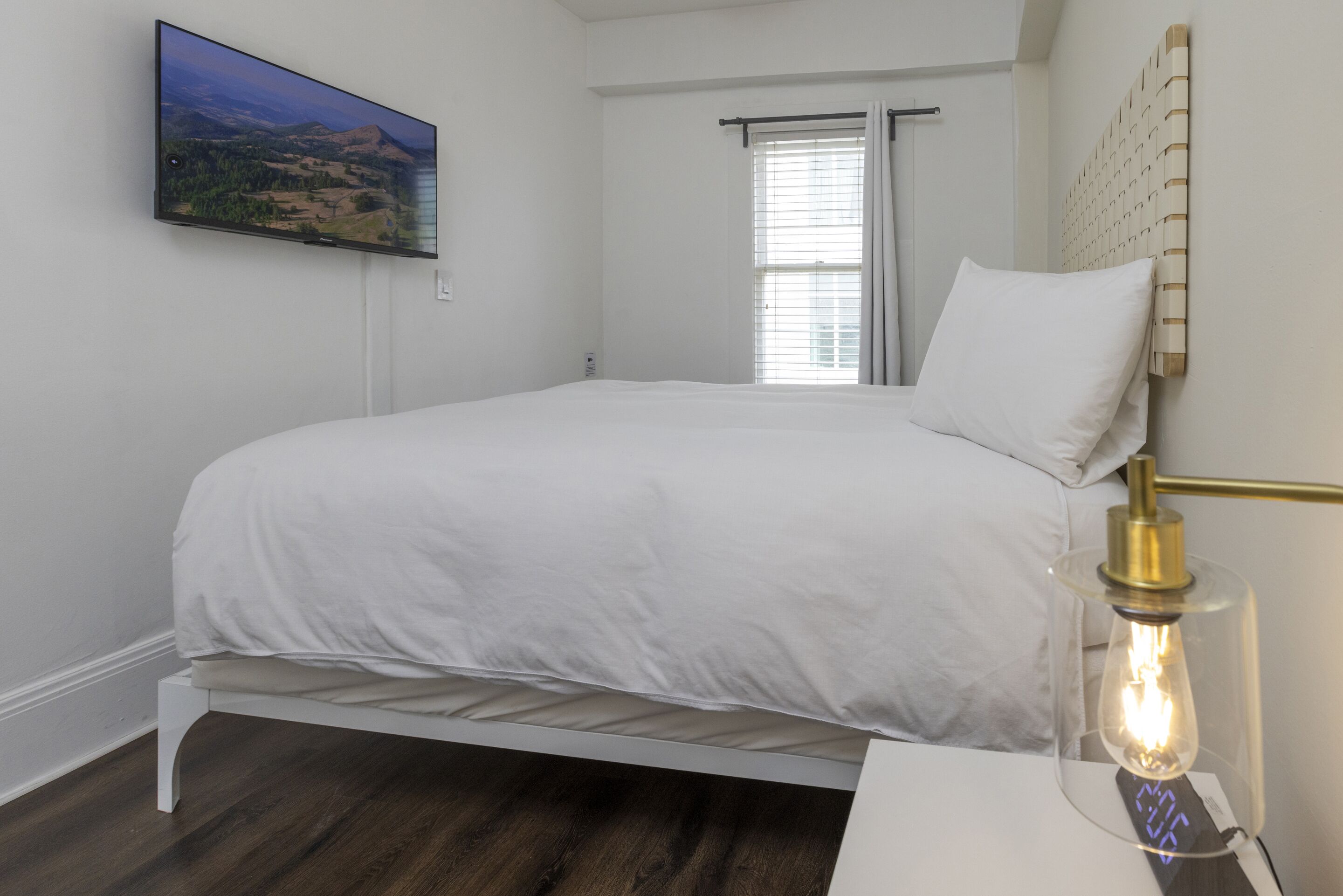 A bedroom with a bed, a mounted TV, a window with curtains, and a bedside table with a lamp.
