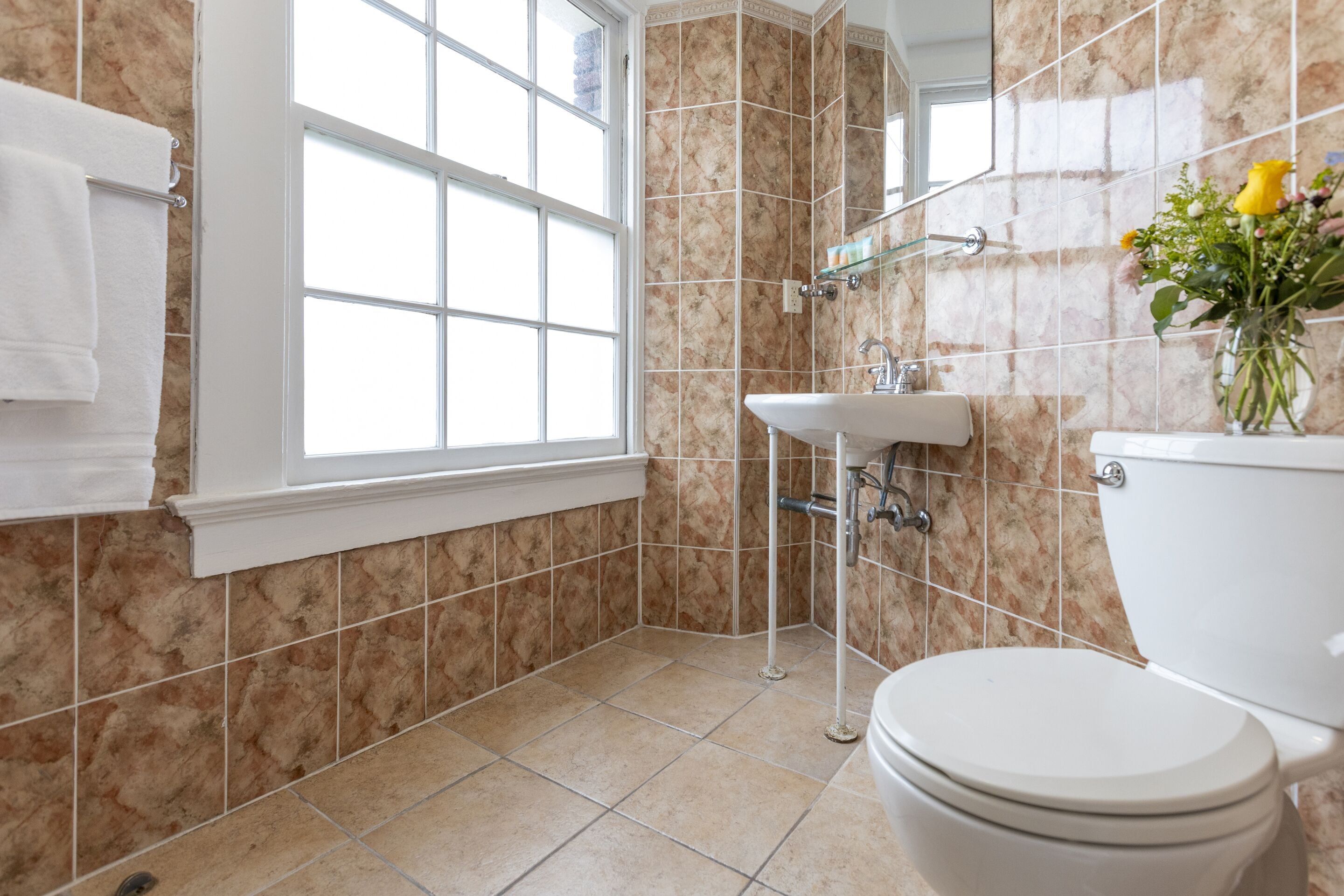 Casa de banho com sanita, lavatório e zona de duche, com azulejos bege e uma janela.