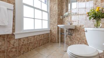 Casa de banho com sanita, lavatório e zona de duche, com azulejos bege e uma janela.