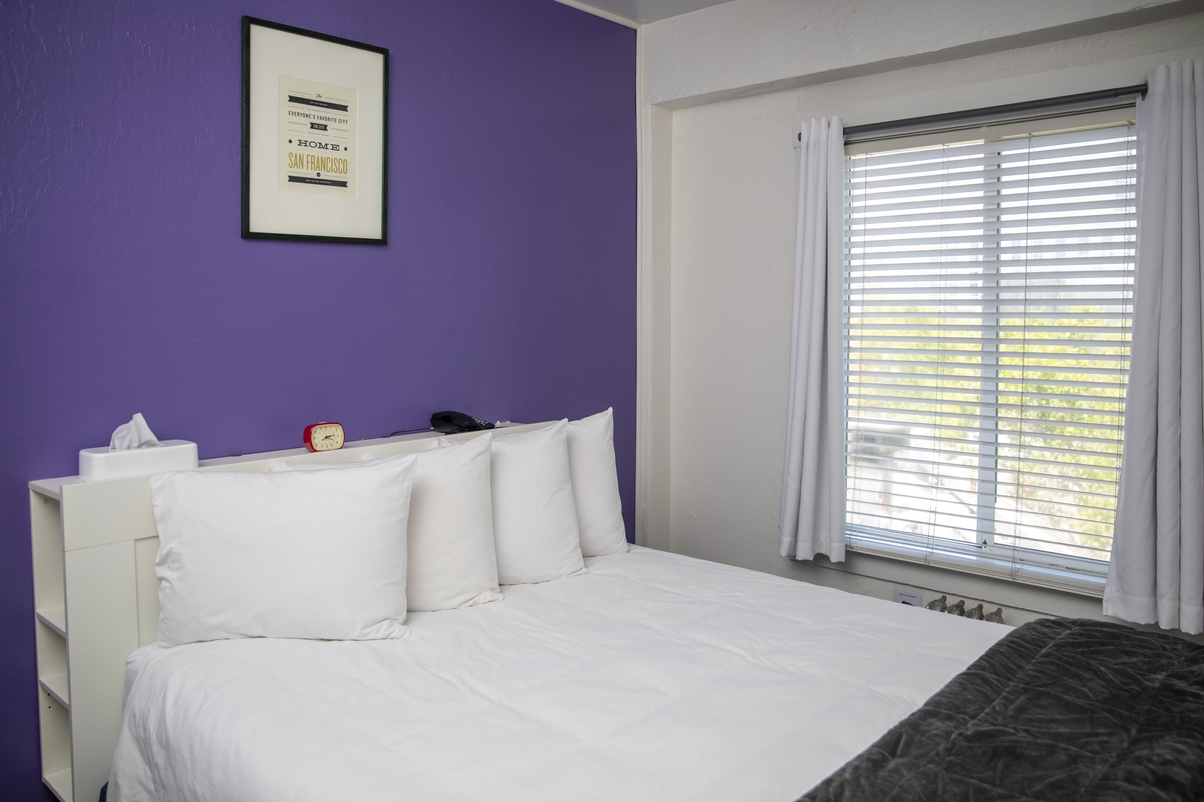 A hotel room with a bed, white headboard, a framed picture on the wall, a bedside table with a tissue box and a red alarm clock, and a window with white curtains.