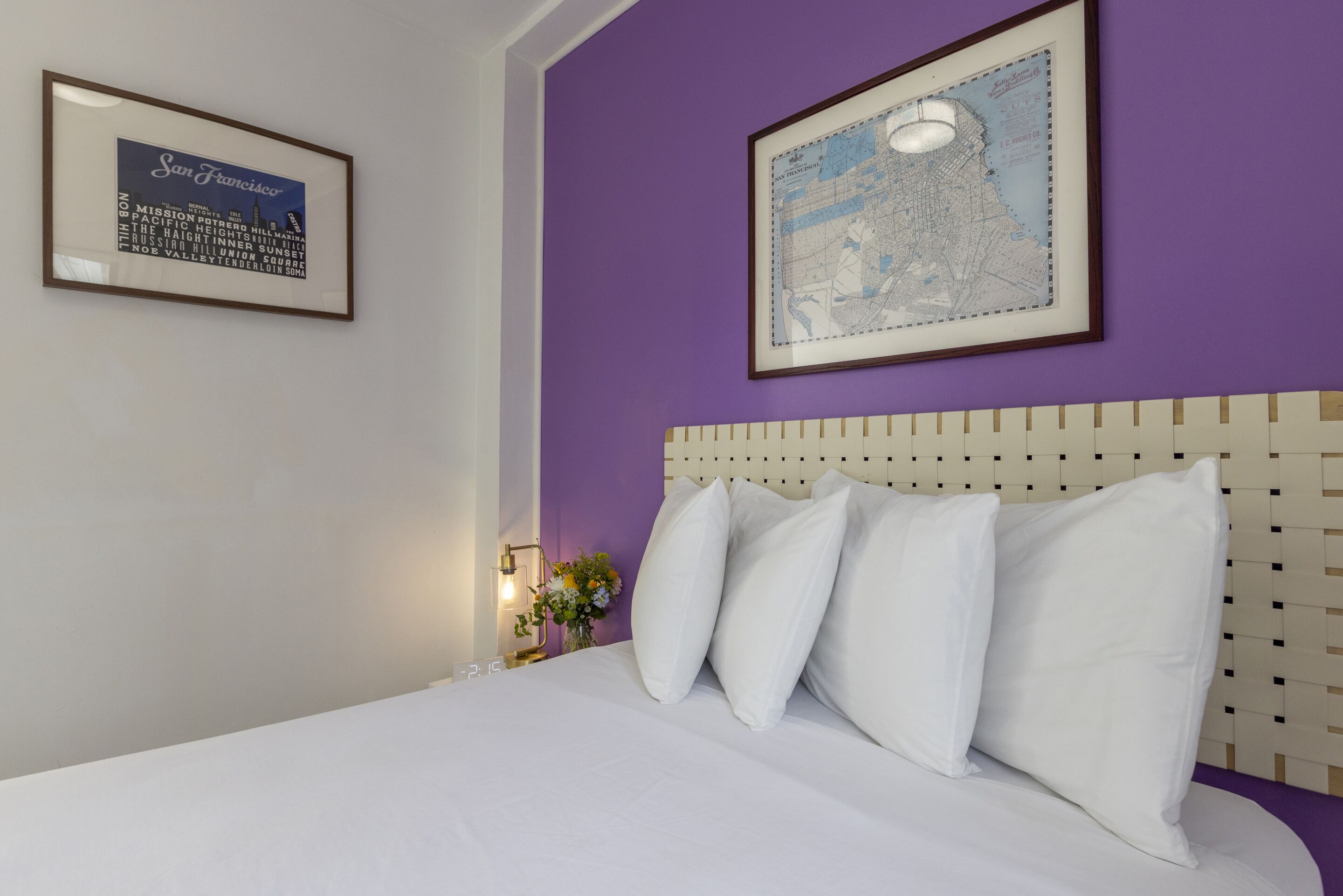 A neatly made bed with white linens and pillows, a purple wall, and two framed pictures, one of a cityscape and the other with text.