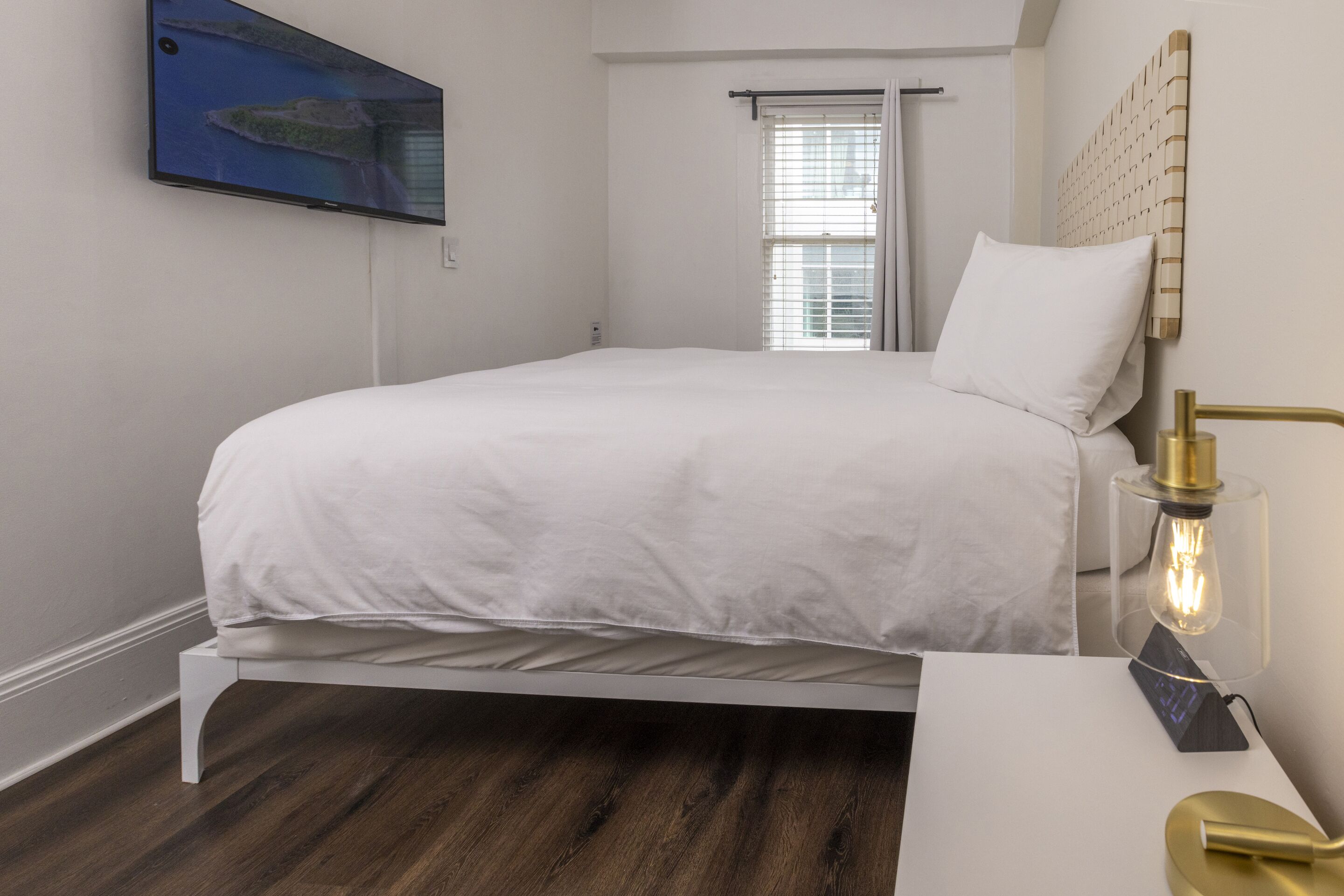 A bedroom with a bed, a TV mounted on the wall, a window with blinds, and a bedside table with a lamp.