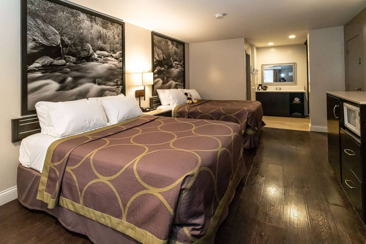 A hotel room with two beds, a large painting of a river, a desk with a lamp, and a kitchenette.