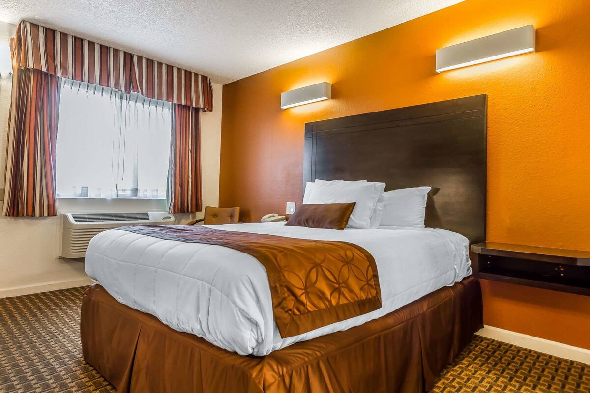 A hotel room with a large bed, a headboard, a window with curtains, an air conditioning unit, and a wall-mounted light fixture.