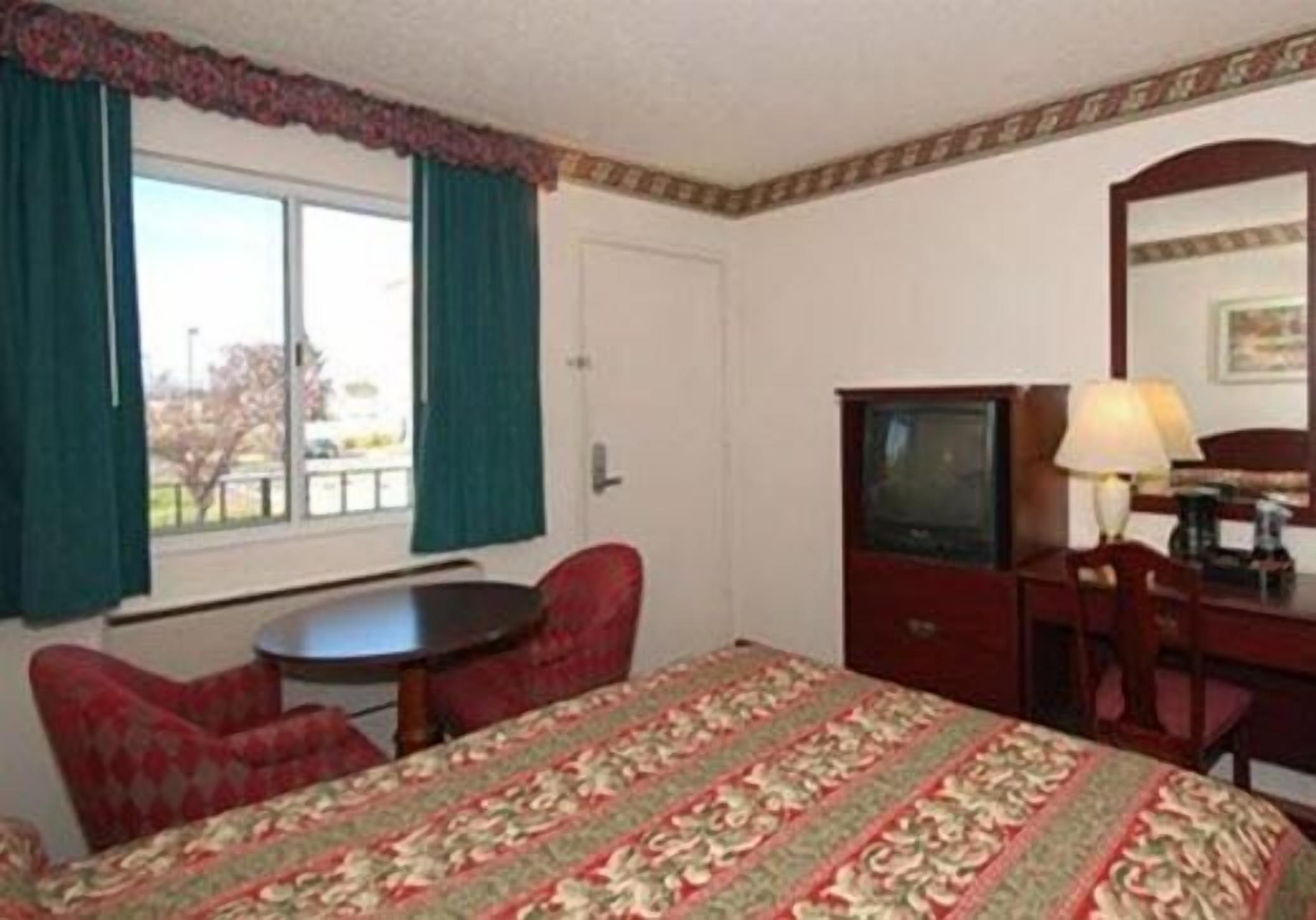 A hotel room with a bed, a desk with a lamp, a television, and a window with curtains.