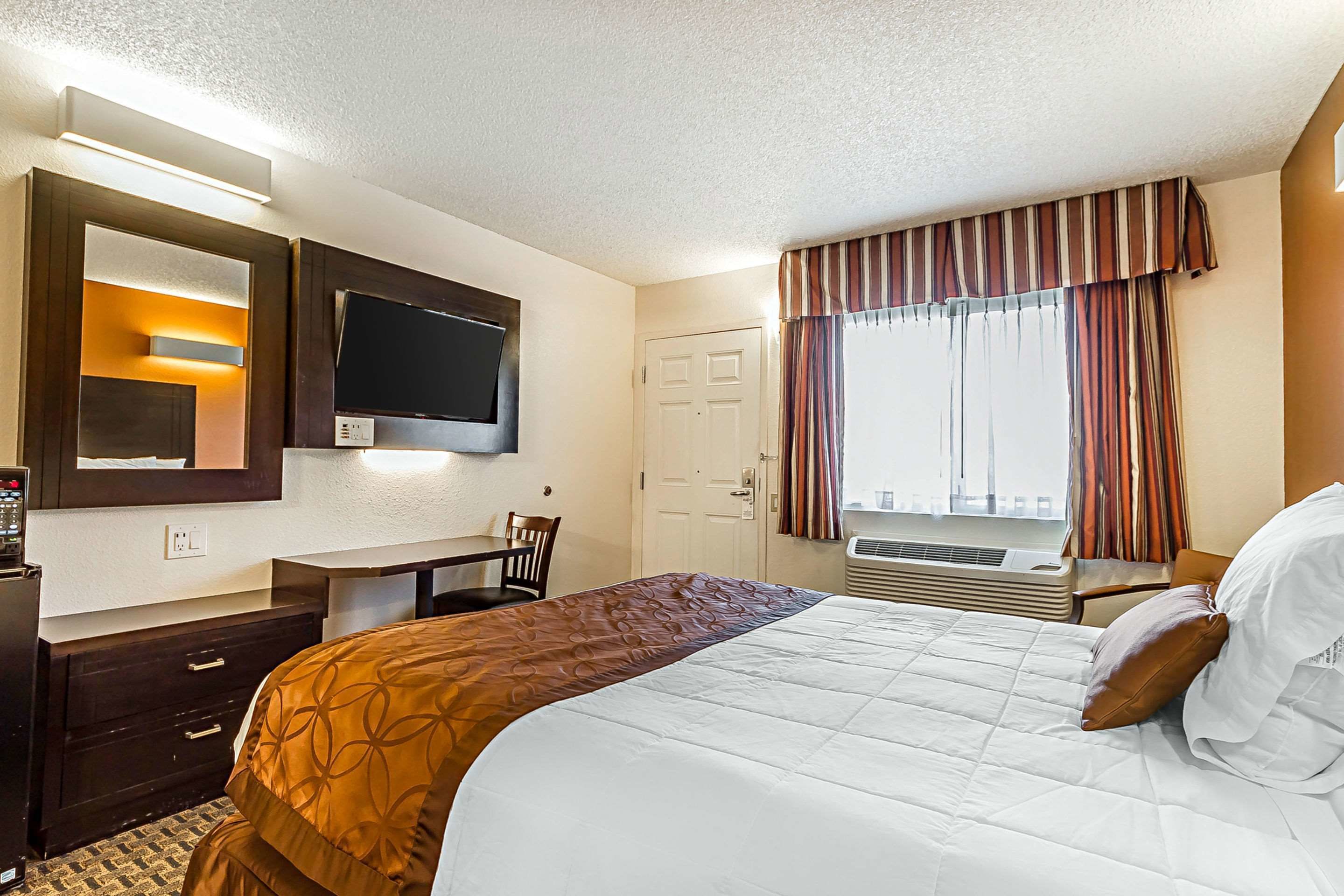 Habitación de hotel con cama, escritorio, televisión, ventana con cortinas y puerta.