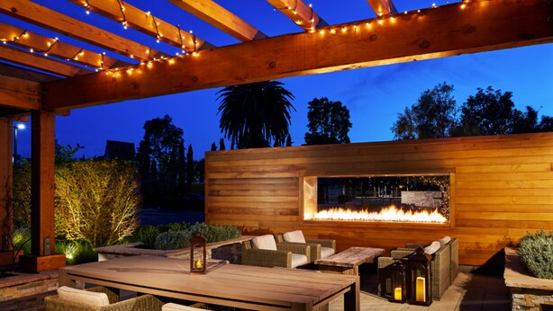 An outdoor patio area with a wooden pergola, wicker furniture, a fire feature, and string lights. - The Nest Hotel Palo Alto (Palo Alto)