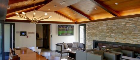 A spacious living area with a wooden ceiling, a stone wall, and a fireplace. There is a dining area with a long table and chairs, and a seating area with sofas and a TV.