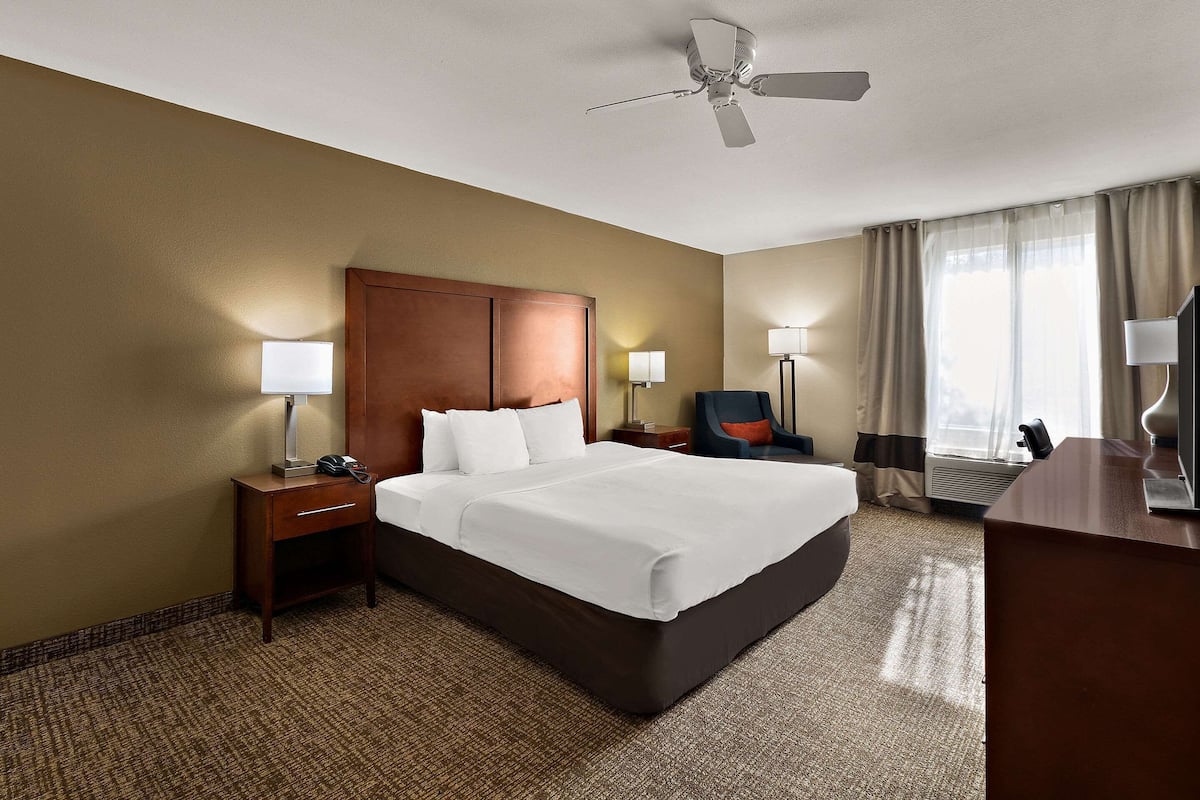 A hotel room with a large bed, a wooden headboard, two bedside tables with lamps, a desk with a chair, and a ceiling fan.