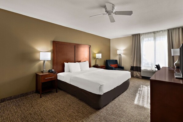 A hotel room with a large bed, a wooden headboard, two bedside tables with lamps, a desk with a chair, and a ceiling fan.
