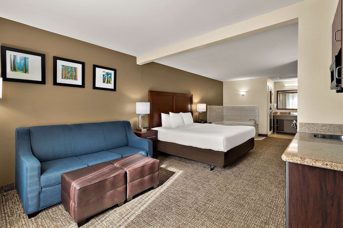 A hotel room with a large bed, a blue sofa, a brown ottoman, and a kitchenette.
