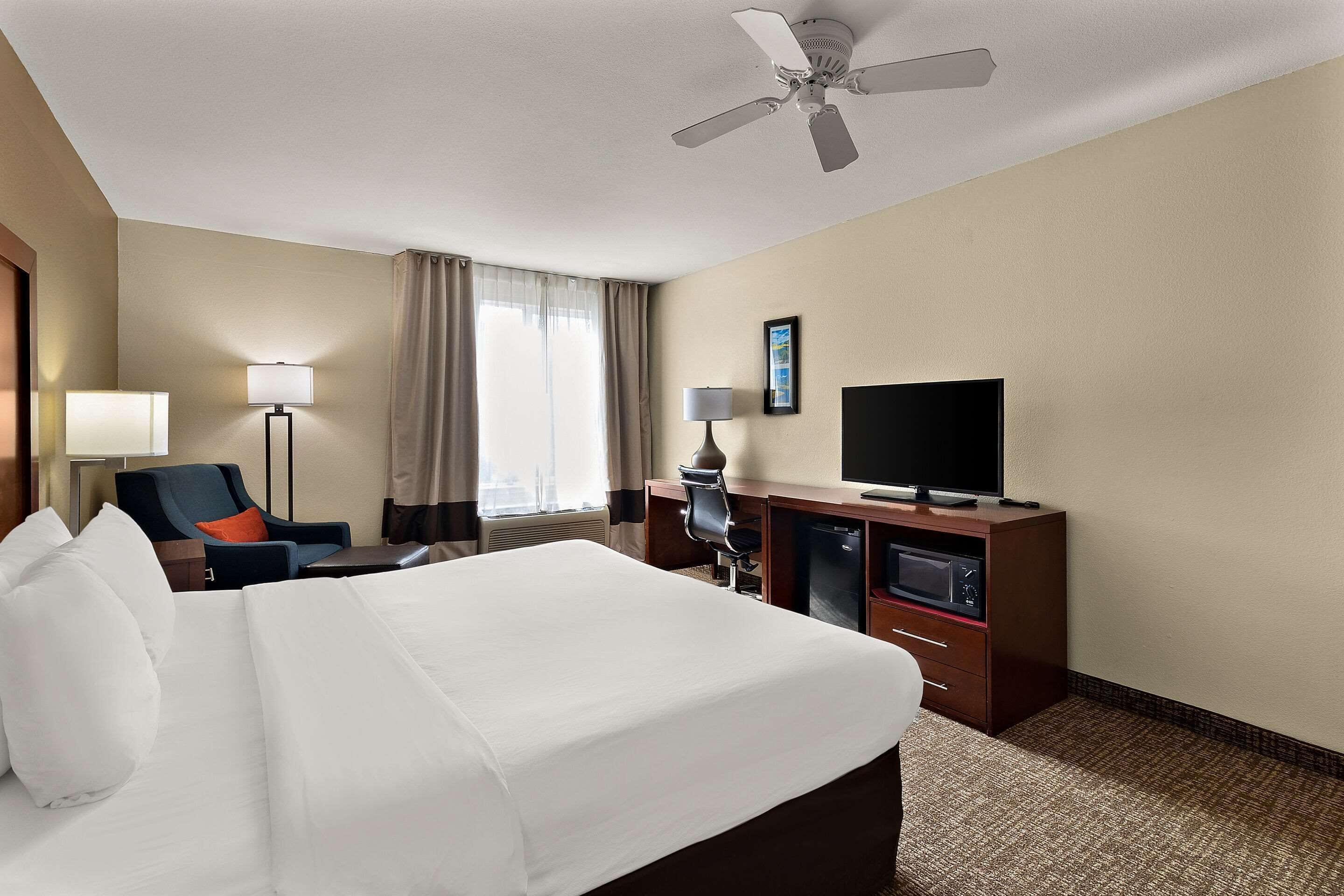 A hotel room with a large bed, a desk with a computer, a television, and a ceiling fan.