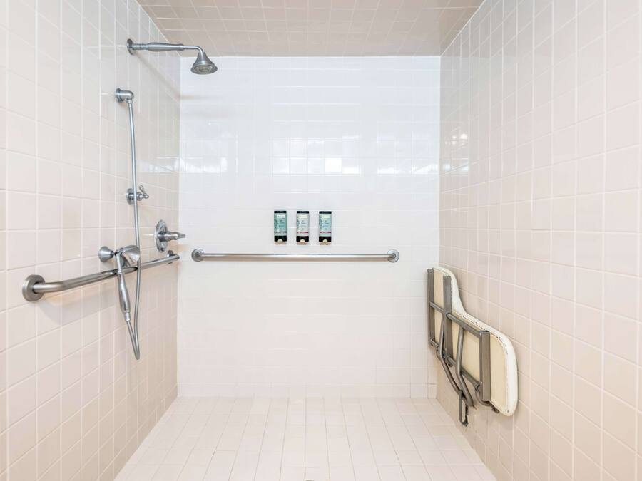 A bathroom with white tiled walls, a shower with a grab bar, and two hand dryers on the wall.